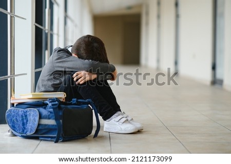 Little boy sitting alone on floor after suffering an act of bullying while children run in the background. Sad young schoolboy sitting on corridor with hands on knees and head between his legs.