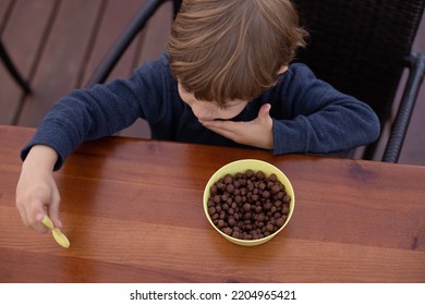 Little Boy Sitting On Chair At Brown Wooden Table In Kitchen And Having Breakfast With Chocolate Balls Top View. Kid Of Kindergarten Age Trifle Spoon In Hand. Ready Breakfast, Not Hungry Child.