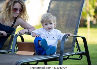 Little Boy Sitting On Deck Chair With His Mom And IPad.