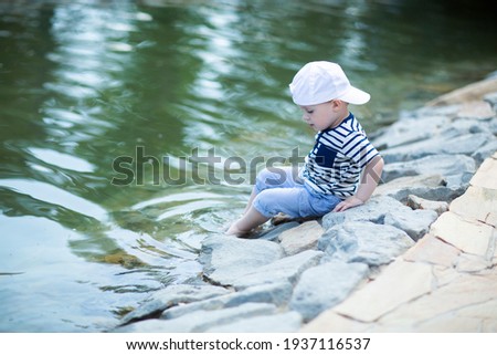 Little boy is sitting on the edge of a pond in a city park and touches the water in the pond with his foot