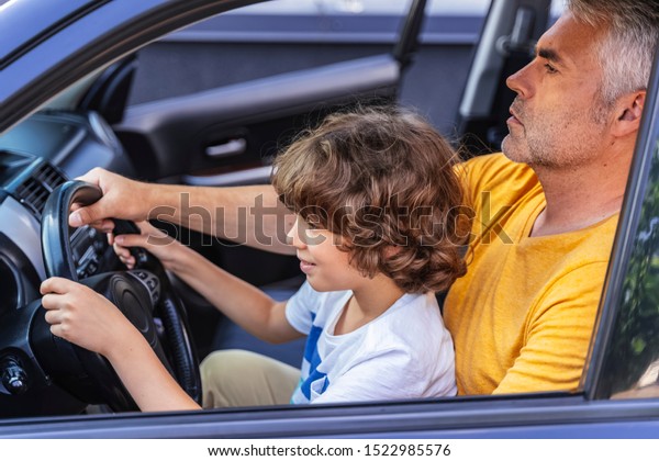 Little boy sitting on knees of his father in the\
car. Driving lessons\
concept