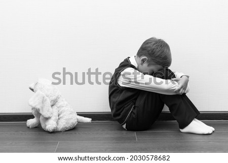 A little boy of six is offended and sits on the floor against a white wall next to a soft toy with his hands closed. Selective focus. Close-up