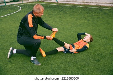 Little Boy, Soccer Player Lying On Grass With Foot Injury. Coach Helps Kid After Getting Injury During Sport Training. Concept Of Health, Assistant, Care, Sport, Competition