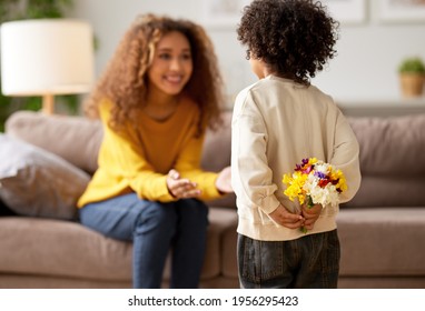 Little Boy Son Holding Present For His Mommy, Hiding Flower Bouquet Behind His Back While Mom Excited African American Race Woman Sitting On Sofa And Waiting, Selective Focus O Child. Mothers Day  