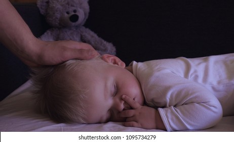 A little boy (son) is sleeping sweetly, blond, mother gently stroking her son, a gray bear toy, in the dark. Concept: children, kids, baby, love, caring family.