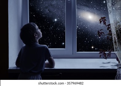 A little boy is standing near the window and looking outside, imagining boundless space with myriad of stars