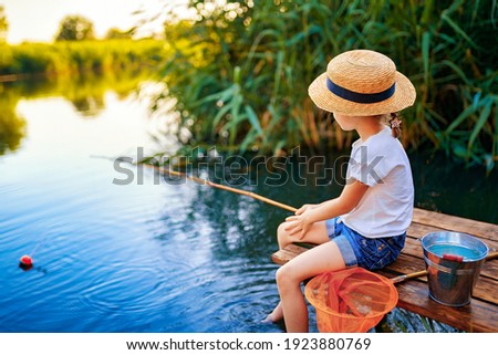 Little boy in straw hat sitting on the edge of a wooden dock and fishing in lake at sunset.