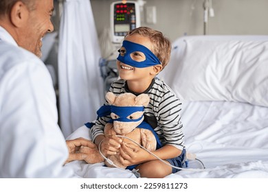 Little boy with superhero mask embracing teddy bear while playing with doctor and sitting at hospital. Playful child dressed as superhero talking to pediatrician. Doctor visiting his little patient. - Powered by Shutterstock