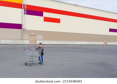 Little Boy With Supermarket Trolley In Front Of Long Warehouse Building