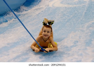 Little Boy Surfing In A Beach Wave Simulator Ride In A Water Amusement Park. A Child Tries To Ride A Bodyboard On Generated Waves.