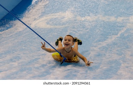 Little Boy Surfing In A Beach Wave Simulator Ride In A Water Amusement Park. A Child Tries To Ride A Bodyboard On Generated Waves.