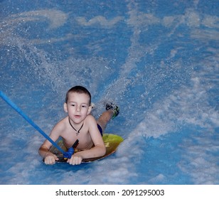 Little Boy Surfing In A Beach Wave Simulator Ride In A Water Amusement Park. A Child Tries To Ride A Bodyboard On Generated Waves.