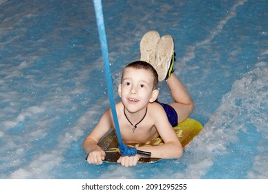 Little Boy Surfing In A Beach Wave Simulator Ride In A Water Amusement Park. A Child Tries To Ride A Bodyboard On Generated Waves.