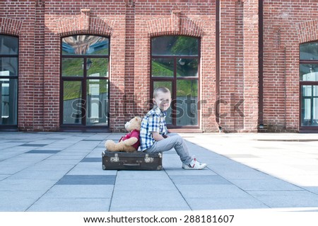 little boy with teddy bear toy is sitting on the suitcase on the deserted street in summer sunny morning