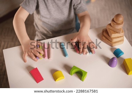 little boy three years old sits at  table with colored crayons and plasticine and plays sensory educational games. Closeup of hands focus on colored plasticine and constructor. Unrecognizable no face