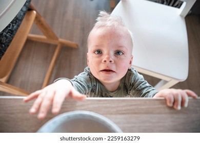 little boy toddler standing in the kitchen at the table reaching for a bowl of porridge, the boy has blue teary eyes tears rolling down his cheeks, stains on his cheeks - Shutterstock ID 2259163279