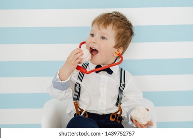 Little Boy With Toy Stethoscope Eating Marshmallow. Adorable Happy Child Playing Doctor While Enjoying Sweet Dessert Medium Shot. Joyful Caucasian Kid Posing On Stripped Wall Background