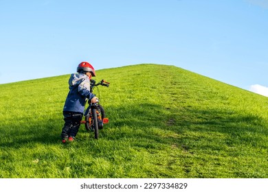 A little boy is trying to climb the hill on his bike. Child development, cycling and children cycling. educational and development activities. Activities in nature. Perseverance and determination. - Shutterstock ID 2297334829