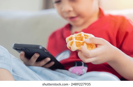 Little boy using phone while eating waffle during lunch or dinner at the home. Nomophobia or Attention Deficit Hyperactivity Disorder ADHD concept.
