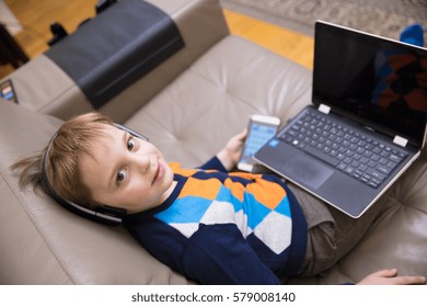 Little boy using a white  laptop computer at home along with mobile phone, wearing headphones, playing some games. - Shutterstock ID 579008140