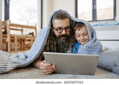 Little boy watching cartoon movie on tablet with father, lying under blanket on floor in kids room. Dad explaining technology to son, digital literacy for kids. - Powered by Shutterstock