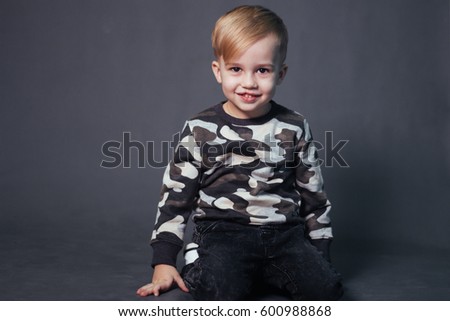 A little boy with white hair and a charming smile. The clothes are military style. Studio photo with a child on a gray background.