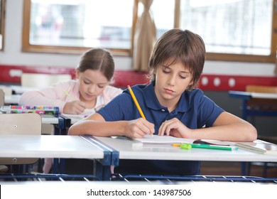 Little boy writing notes while classmate studying in background at classroom