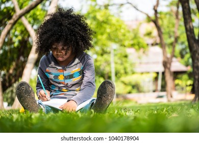 A little boy writing on notebook while sitting on green grass in a park - Powered by Shutterstock