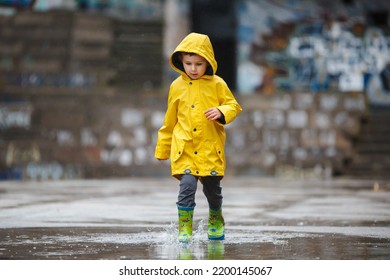 Little boy in a yellow raincoat and green rubber boots runs through a puddle on a gloomy autumn day - Shutterstock ID 2200145067