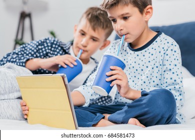 Little Boys Using Tablet And Drinking Soda While Lying In Bed At Home