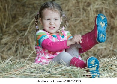 Little Braided Girl Putting On Red Gum Boots On Dried Loose Grass Hay In Farm Haystack