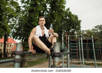 Little Break. Young Athletic Guy With Muscular Body, Sportsman And Fitness Model, Strong Man Sitting On Parallel Bars. Athletic Body Shape, Guy Relax At Outdoor Gym.