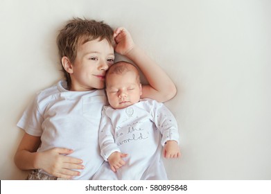 Little brother hugging her newborn baby. Toddler kid meeting new sibling. Cute boy and new born baby girl relax in a white bedroom. Family with children at home. Love, trust and tenderness