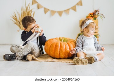 Little Brother And Sister Are Sitting On The Floor Next To A Large Pumpkin. Children's Photo Zone In Autumn Style With Pumpkins. The Boy Takes Pictures Of His Sister With A Toy Camera.