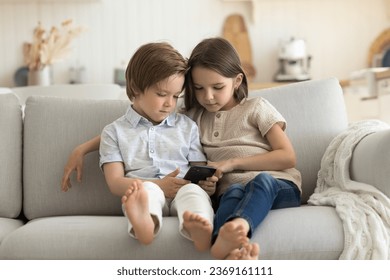 Little brother and sister spend time together with smartphone sitting on couch, watch educational video, addicted to internet, enjoy leisure online without parent. Younger generation use modern tech - Powered by Shutterstock