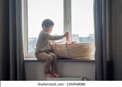 Little brother is sitting near the window with himnewborn sister in the cradle. Children with small age difference. Happy Family concept.