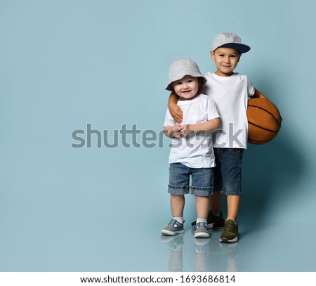 Little brunet boy hugging his toddler brother or sister. They dressed in casual clothes. Elder one holding basketball ball, smiling posing on blue background. Childhood, sport. Full length, copy space