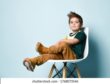 Little brunet child in colorful t-shirt, brown pants and sneakers. Smiling and looking at you while sitting on white chair against blue studio background. 