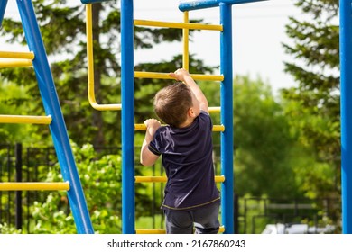 A Little Caucasian Athletic Boy Climbs On The Horizontal Bars On The Playground. A Sporty Child Does Exercises On The Monkey Bars In Kindergarten. Sports And Activities. Healthy Lifestyle