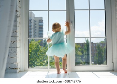 Little Caucasian Baby Girl At Home In White Room Stands Near Window, Watching Outside