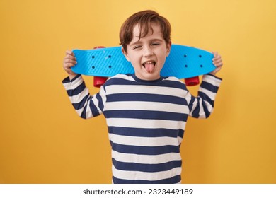 Little caucasian boy holding skate over yellow background sticking tongue out happy with funny expression.  - Shutterstock ID 2323449189
