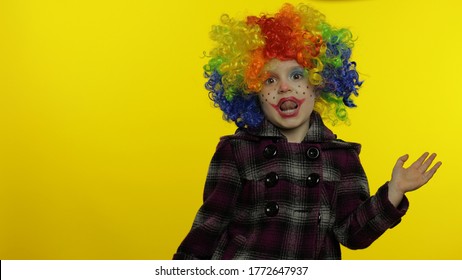 Little caucasian child girl clown in rainbow wig making silly faces. Five years old female kid having fun, smiling, dancing, looking at camera. Expressions. Copy space. Halloween. Yellow background