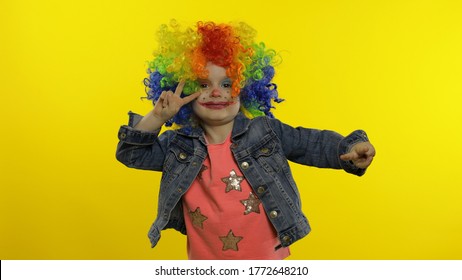 Little caucasian child girl in clown cosplay making silly faces. Colorful wig. Happy five years old female kid having fun, smiling, dancing, looking at camera. Halloween. Isolated on yellow background