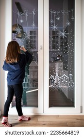 Little caucasian child girl decorating home window for Christmas and white pencil (liquid chalk)  Festive home decor idea  Vertical shot 