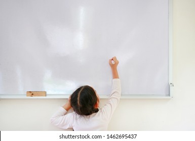 Little caucasian child girl writing something on whiteboard with a marker in the classroom. White board with copy space for text. Back view.