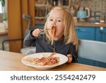 Little Caucasian child toddler girl eats spaghetti with tomato sauce at home with a fork.