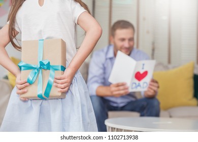 Little Caucasian Girl Daughter Congratulate Daddy Hiding Giving A Surprise Card And Gift Box. Family Holiday Father And Girl Celebrate Together. Love Lifestyle Happy Father’s Day Holiday Party Concept