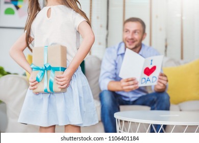 Little Caucasian Girl Daughter Congratulate Daddy Hiding Giving A Surprise Card And Gift Box. Family Holiday Father And Girl Celebrate Together. Love Lifestyle Happy Father’s Day Party Concept