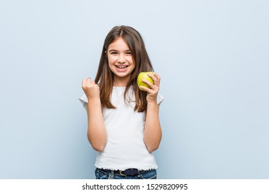 Little caucasian girl holding a green apple cheering carefree and excited. Victory concept.