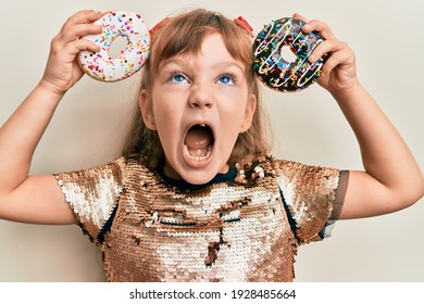 Little Caucasian Girl Kid Holding Tasty Colorful Doughnuts Angry And Mad Screaming Frustrated And Furious, Shouting With Anger Looking Up. 